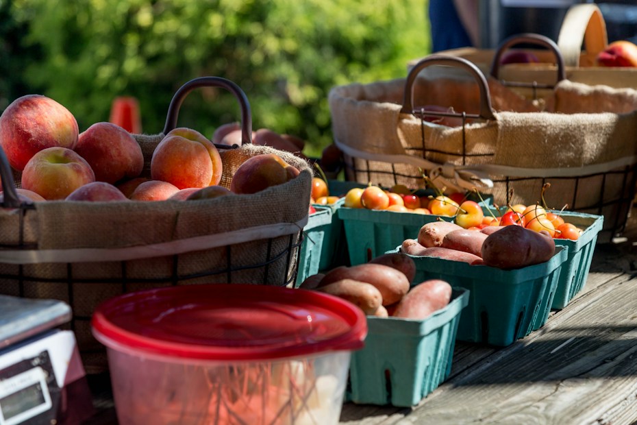 Baskets filled with ripe peaches and small boxes containing potatoes and colorful cherry tomatoes are arranged on a wooden table. The sunlight casts gentle shadows, enhancing the fresh and natural look of the produce. A red container with a lid is partially visible in the foreground.