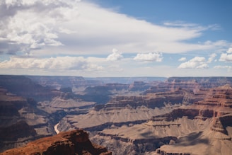 A vast landscape featuring the majestic Grand Canyon. The image captures the intricate layers of rock formations, with shades ranging from deep reds to soft browns. The canyon's vastness is emphasized by the river cutting through the base, and the expansive sky above is dotted with fluffy white clouds.