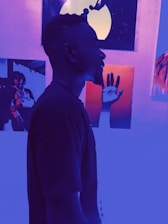 A person is standing in profile, silhouetted against a vibrant background featuring various artworks. The lighting casts a blue and purple hue across the scene, creating a dramatic contrast with the colorful art pieces. The artworks include a hand reaching up and an abstract design with an orange and black color scheme.