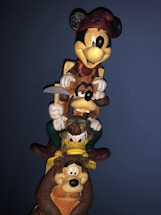A stack of cartoon character sculptures including a mouse wearing a hat and overalls holding an axe, a dog with large ears, a duck with a yellow bill, and a bear at the bottom.
