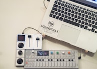 A laptop with the logo 'Soundtrax Stock Music Library' next to a music mixing console and a synthesizer placed on a light-colored surface. The synthesizer has colorful knobs and a small display screen.