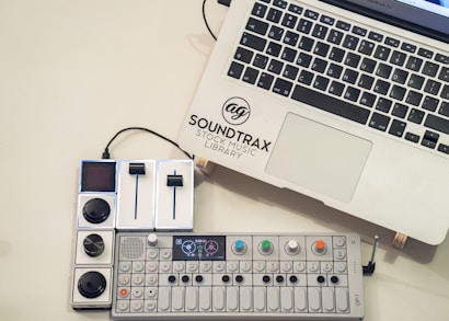 A laptop with the logo 'Soundtrax Stock Music Library' next to a music mixing console and a synthesizer placed on a light-colored surface. The synthesizer has colorful knobs and a small display screen.