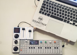 A laptop with the logo 'Soundtrax Stock Music Library' next to a music mixing console and a synthesizer placed on a light-colored surface. The synthesizer has colorful knobs and a small display screen.