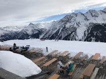A snowy mountain landscape with wooden benches and tables covered in snow. People are sitting at the tables, enjoying the view. In the background, towering snow-covered mountains are visible under a partially cloudy sky.
