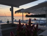 A serene beachside view at sunset, featuring an outdoor seating area with lounge chairs and sun umbrellas. In the foreground, there are pink flowers on a counter. The horizon shows a peaceful sea, while the sun sets behind a distant hill, casting warm orange and pink hues across the sky.
