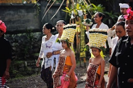 A group of people is participating in a traditional cultural procession. Two young children are dressed in vibrant traditional attire with elaborate floral headdresses, leading the group. The older participants, wearing white lace garments, accompany them. The setting appears to be an outdoor area with foliage in the background and a stone wall.