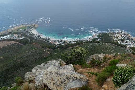 A coastal landscape with a scenic view of the ocean and shoreline. The area is dotted with numerous buildings, likely residential, nestled into the green hills. The water displays a range of blue hues, with waves crashing against the shore. Vegetation appears lush on the hillsides, and large rocks are visible in the foreground.