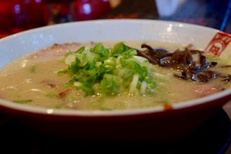 A close-up of a bowl of ramen soup garnished with chopped green onions and dark leafy vegetables. The broth is creamy and features slices of meat submerged beneath the surface.