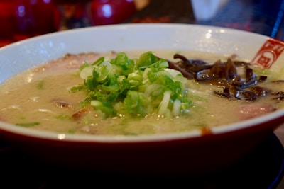 A close-up of a bowl of ramen soup garnished with chopped green onions and dark leafy vegetables. The broth is creamy and features slices of meat submerged beneath the surface.