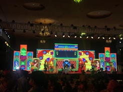A lively stage performance is in progress with performers dressed in traditional attire. They are holding colorful umbrellas, dancing against a backdrop of vibrant, illuminated screens displaying intricate patterns and text. The venue has a decorative ceiling with chandeliers and a grid of stage lights.
