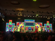 A lively stage performance is in progress with performers dressed in traditional attire. They are holding colorful umbrellas, dancing against a backdrop of vibrant, illuminated screens displaying intricate patterns and text. The venue has a decorative ceiling with chandeliers and a grid of stage lights.