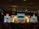 A lively stage performance is in progress with performers dressed in traditional attire. They are holding colorful umbrellas, dancing against a backdrop of vibrant, illuminated screens displaying intricate patterns and text. The venue has a decorative ceiling with chandeliers and a grid of stage lights.