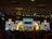 A lively stage performance is in progress with performers dressed in traditional attire. They are holding colorful umbrellas, dancing against a backdrop of vibrant, illuminated screens displaying intricate patterns and text. The venue has a decorative ceiling with chandeliers and a grid of stage lights.