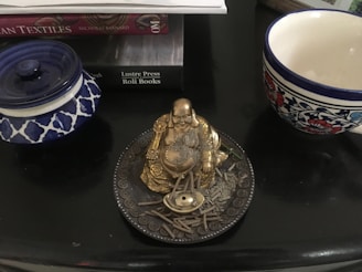 A golden laughing Buddha figurine is placed on a circular plate with an ornamental design, surrounded by ashes and incense sticks. On the left, there is a blue and white patterned container with a lid. To the right, there is a cup with a floral design featuring blue and red colors. Behind these items, a few books are stacked, and one book partially titled 'Indian Textiles' is visible.