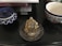 A golden laughing Buddha figurine is placed on a circular plate with an ornamental design, surrounded by ashes and incense sticks. On the left, there is a blue and white patterned container with a lid. To the right, there is a cup with a floral design featuring blue and red colors. Behind these items, a few books are stacked, and one book partially titled 'Indian Textiles' is visible.