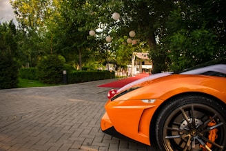 An orange sports car is parked on a cobblestone driveway surrounded by lush green trees and bushes. White lanterns hang from the trees, creating a festive atmosphere. In the background, a red carpet leads to an elegant tent or pavilion, suggesting a special event or celebration.