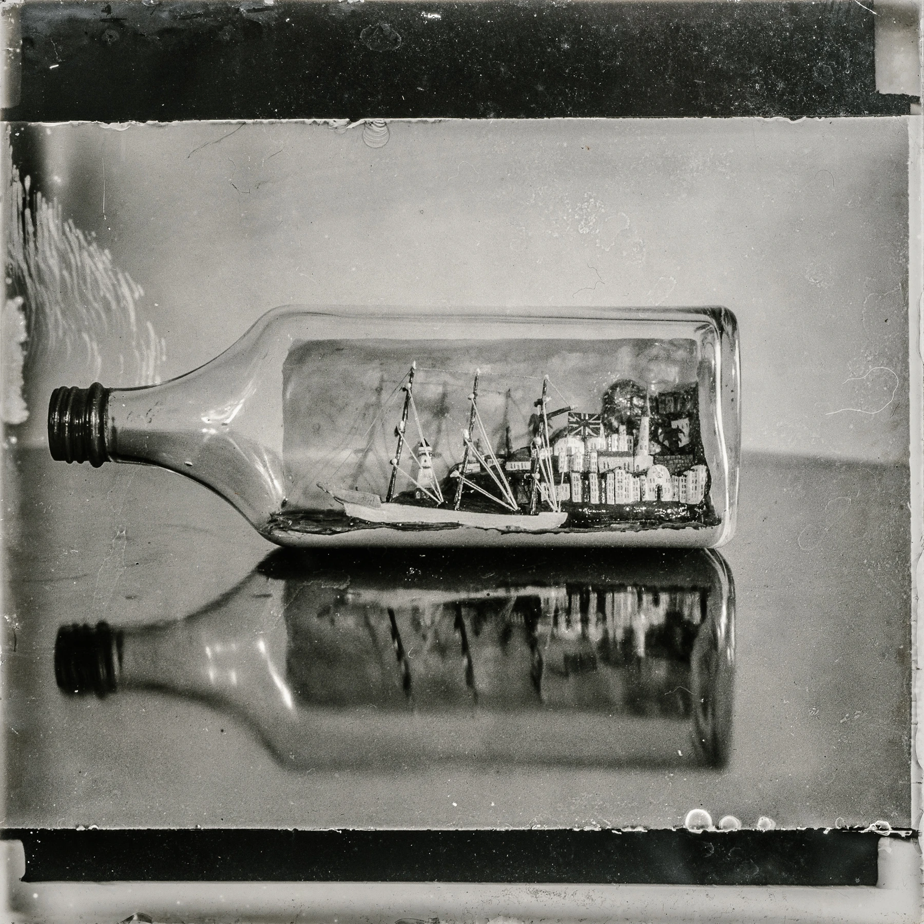Black and white photo of a ship inside a flat-sided glass bottle lying on a polished table, mirrored in the surface