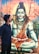 A young man stands looking at a large, detailed painting of a deity with blue skin, wearing a garland of rudraksha beads and a snake around its neck. The deity appears to be sitting in a meditative pose on a tiger skin, with mountains and a trishula (trident) in the background.