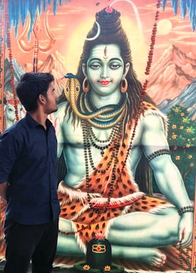 A young man stands looking at a large, detailed painting of a deity with blue skin, wearing a garland of rudraksha beads and a snake around its neck. The deity appears to be sitting in a meditative pose on a tiger skin, with mountains and a trishula (trident) in the background.