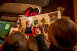 A child is holding a colorful picture book with illustrations of people in vibrant outfits. Two sets of hands are visible touching the pages, indicating engagement with the story. The background includes a pair of pink and black sneakers on a brown surface.