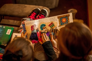 A child is holding a colorful picture book with illustrations of people in vibrant outfits. Two sets of hands are visible touching the pages, indicating engagement with the story. The background includes a pair of pink and black sneakers on a brown surface.