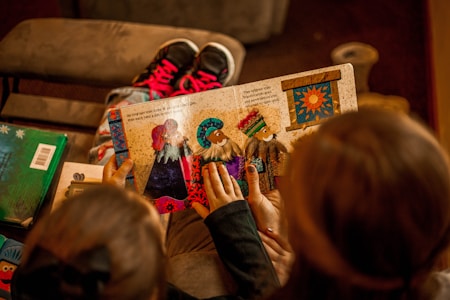 A child is holding a colorful picture book with illustrations of people in vibrant outfits. Two sets of hands are visible touching the pages, indicating engagement with the story. The background includes a pair of pink and black sneakers on a brown surface.