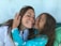 A woman and a child are sharing a joyful moment with smiles. The child is playfully holding the woman's face, and both have their eyes closed. Hair is slightly messy, suggesting movement or a breeze.