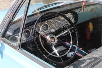 The interior of a vintage car can be seen with a focus on the dashboard and steering wheel. The steering wheel has a classic design with an emblem in the center, surrounded by retro-style gauges on the dashboard. A small hanging decoration is visible near the rearview mirror. Sunlight casts shadows on the interior, revealing a mix of small items like a fire extinguisher and various personal belongings on the seat.