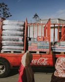 A large red Coca-Cola truck loaded with stacks of plastic-wrapped soda bottles is parked in front of a building with decorative architecture. Two people, partially visible, walk by in the foreground. The sky is clear with a few scattered clouds, and trees with sparse leaves are visible.