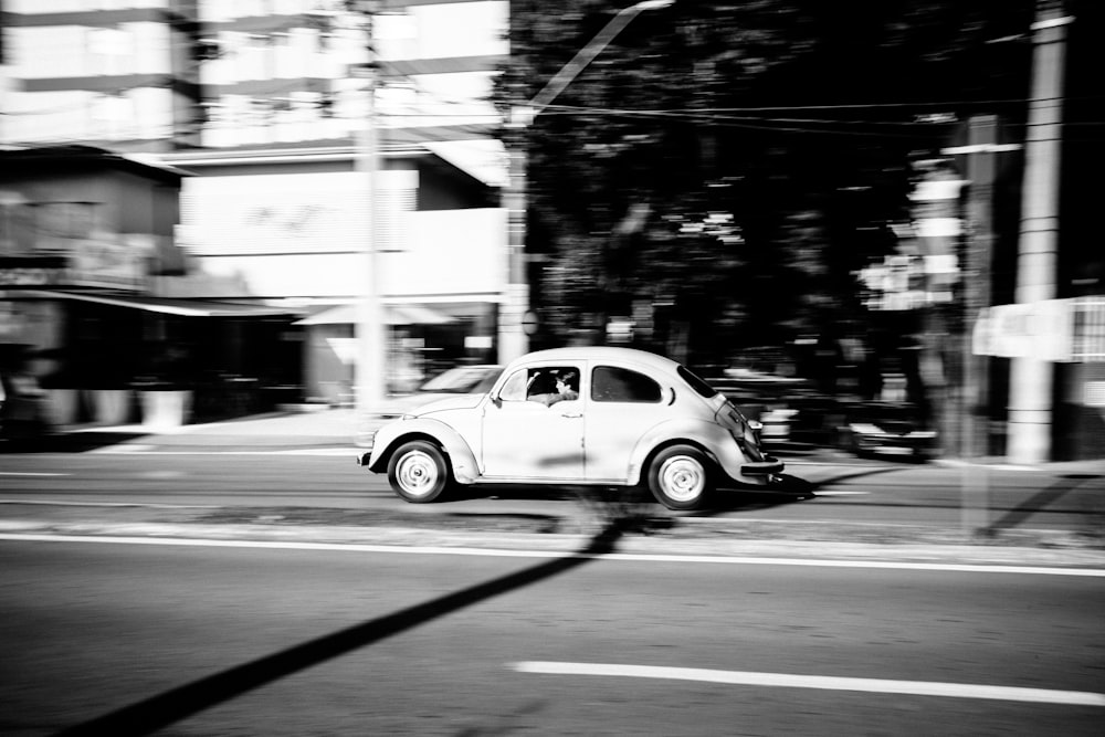 photo en niveaux de gris d’une voiture Volkswagen Coccinelle blanche roulant sur une route grise