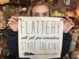 A person holds a sign over their face in a shop filled with decorative items. The sign reads 'Flattery will get you somewhere, start talking' and the person has sunglasses resting on their head.