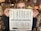 A person holds a sign over their face in a shop filled with decorative items. The sign reads 'Flattery will get you somewhere, start talking' and the person has sunglasses resting on their head.