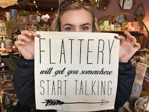 A person holds a sign over their face in a shop filled with decorative items. The sign reads 'Flattery will get you somewhere, start talking' and the person has sunglasses resting on their head.