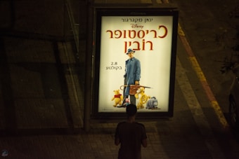 An outdoor advertisement for a movie featuring characters from a well-known children's story, including a man in a blue coat and hat, and animated animals like a bear and a donkey. The ad is brightly lit and placed on a dimly lit sidewalk with a person standing in front of it.