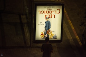 An outdoor advertisement for a movie featuring characters from a well-known children's story, including a man in a blue coat and hat, and animated animals like a bear and a donkey. The ad is brightly lit and placed on a dimly lit sidewalk with a person standing in front of it.