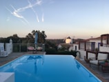 A serene outdoor scene featuring a clear blue swimming pool with lounge chairs and an umbrella by the poolside. In the background, there is a view of a traditional windmill and several charming white buildings with red roofs. The setting is surrounded by lush greenery and hills under a clear sky with a few contrails visible.