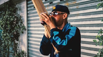 A person wearing sunglasses and a cap plays a wind instrument made of bamboo reeds. They are standing next to a metal shutter with some foliage visible on the side. The individual is dressed in a dark jacket with blue accents.