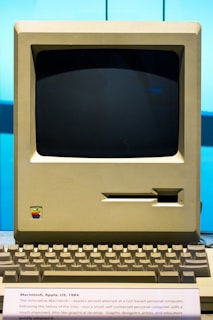 A vintage Apple Macintosh computer from 1984 featuring a boxy monitor with a black screen, beige casing, and an integrated floppy disk drive slot. Below the monitor is a keyboard with similar beige keys. The iconic rainbow Apple logo is present on the casing. The display is set in a museum-like environment with an informational plaque in front.
