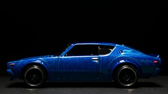 A model of a vintage car with a sleek design in metallic blue, displayed against a dark background. The car features smooth curves and intricate detailing on the body.