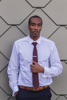 A person wearing a white dress shirt, maroon tie, and brown belt stands against a geometric, taupe-colored wall. The individual has a neutral expression and a well-groomed appearance.