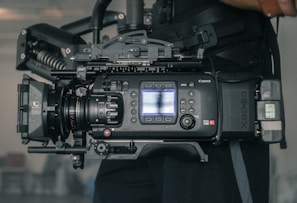 A professional digital video camera with various controls and buttons, mounted on a stabilizing rig. The camera features a lens with focus and zoom rings, as well as a small display screen on the side. The setup appears to be robust and suited for high-quality video production.