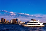 A large white yacht sails across a body of water against the backdrop of a city skyline with multiple high-rise buildings. Fluffy clouds are scattered across a bright blue sky.