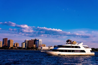 A large white yacht sails across a body of water against the backdrop of a city skyline with multiple high-rise buildings. Fluffy clouds are scattered across a bright blue sky.