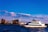 A large white yacht sails across a body of water against the backdrop of a city skyline with multiple high-rise buildings. Fluffy clouds are scattered across a bright blue sky.