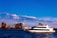 A large white yacht sails across a body of water against the backdrop of a city skyline with multiple high-rise buildings. Fluffy clouds are scattered across a bright blue sky.