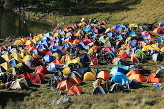A colorful array of numerous tents is set up on a grassy field near a body of water, with people moving around the encampment. The scene conveys the hustle and bustle of a camping event or festival, with vibrant colors and a lively atmosphere. The tents' arrangement and the natural surroundings suggest a harmonious blend of outdoor adventure and social gathering.