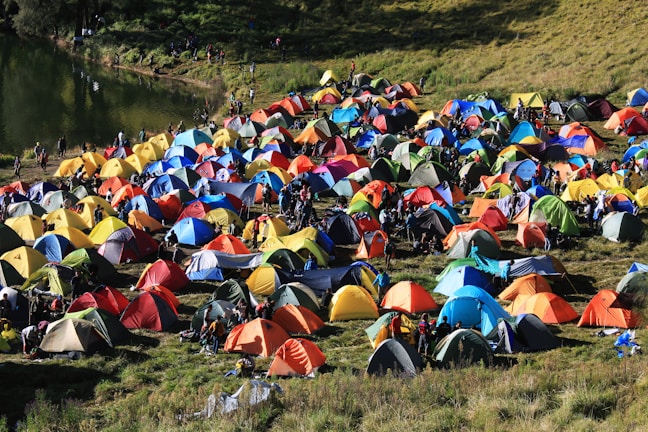 A colorful array of numerous tents is set up on a grassy field near a body of water, with people moving around the encampment. The scene conveys the hustle and bustle of a camping event or festival, with vibrant colors and a lively atmosphere. The tents' arrangement and the natural surroundings suggest a harmonious blend of outdoor adventure and social gathering.