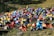 A colorful array of numerous tents is set up on a grassy field near a body of water, with people moving around the encampment. The scene conveys the hustle and bustle of a camping event or festival, with vibrant colors and a lively atmosphere. The tents' arrangement and the natural surroundings suggest a harmonious blend of outdoor adventure and social gathering.