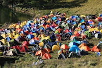 A colorful array of numerous tents is set up on a grassy field near a body of water, with people moving around the encampment. The scene conveys the hustle and bustle of a camping event or festival, with vibrant colors and a lively atmosphere. The tents' arrangement and the natural surroundings suggest a harmonious blend of outdoor adventure and social gathering.