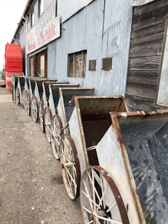 A row of old, metal carts with large, rusted wheels is positioned along the side of a weathered building. The building has a mix of corrugated metal and wooden siding, with signs and small boarded-up windows. In the background, there are red plastic seats stacked on the pavement.